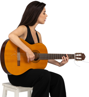 Вид в три четверти сидящей молодой леди в черном костюме, играющей на гитаре