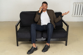 Front view of a perplexed young man sitting on a sofa and talking on his phone