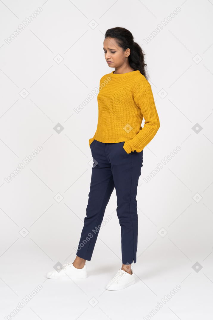 Vista frontal de uma garota chateada em roupas casuais, posando com as mãos nos bolsos e olhando para baixo
