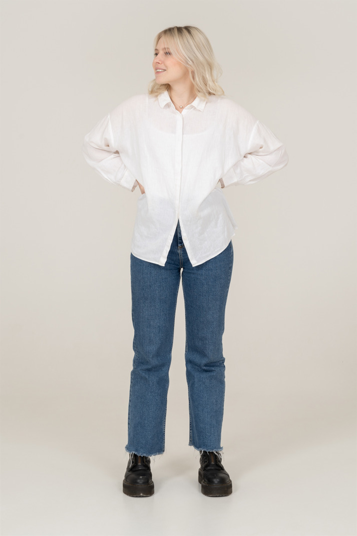 Front view of a blonde female in casual clothes putting hands on hips and looking aside while smiling