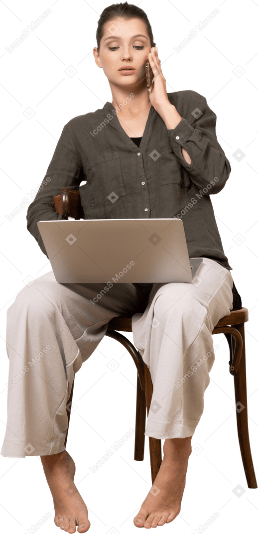 Vorderansicht einer beschäftigten jungen frau, die mit einem laptop und einem handy auf einem stuhl sitzt