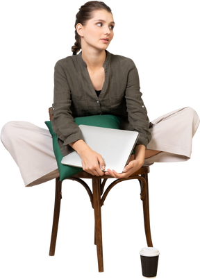 Front view of a bored young woman sitting on a chair and holding her laptop