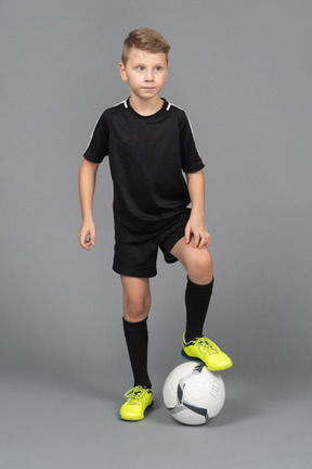 Vista frontal de un niño en uniforme de fútbol poniendo su pie en la pelota y mirando a un lado