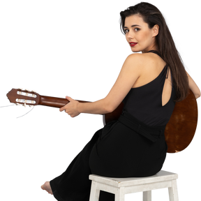 Vista traseira de uma jovem sentada de terno preto segurando o violão enquanto se vira