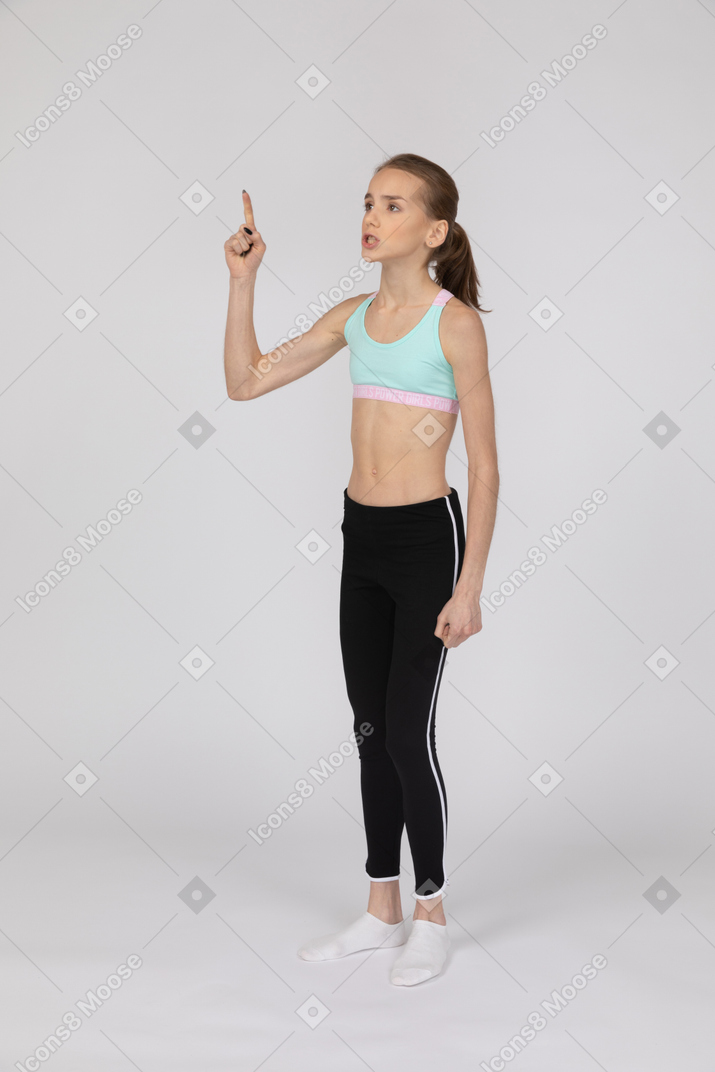 Vista de tres cuartos de una jovencita en ropa deportiva apuntando con el dedo y mirando hacia arriba