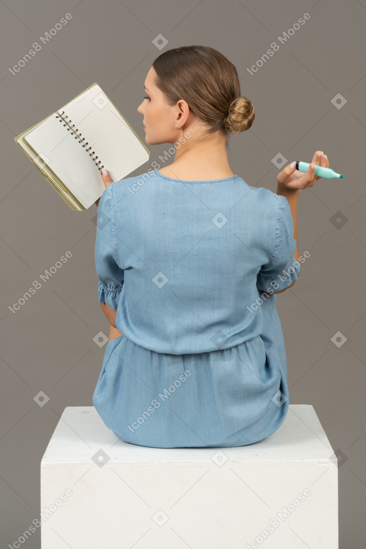 Back view of young woman in blue dress sitting on a cube and making notes