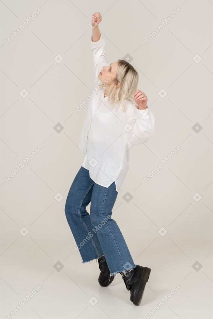 Three-quarter view of a blonde female in casual clothes dancing on her tiptoes and raising hands and head