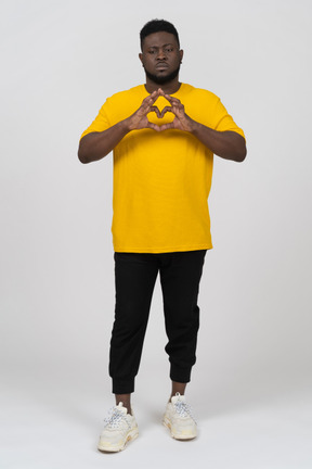 Vista frontale di un cupo giovane uomo dalla pelle scura in maglietta gialla che mostra il gesto del cuore