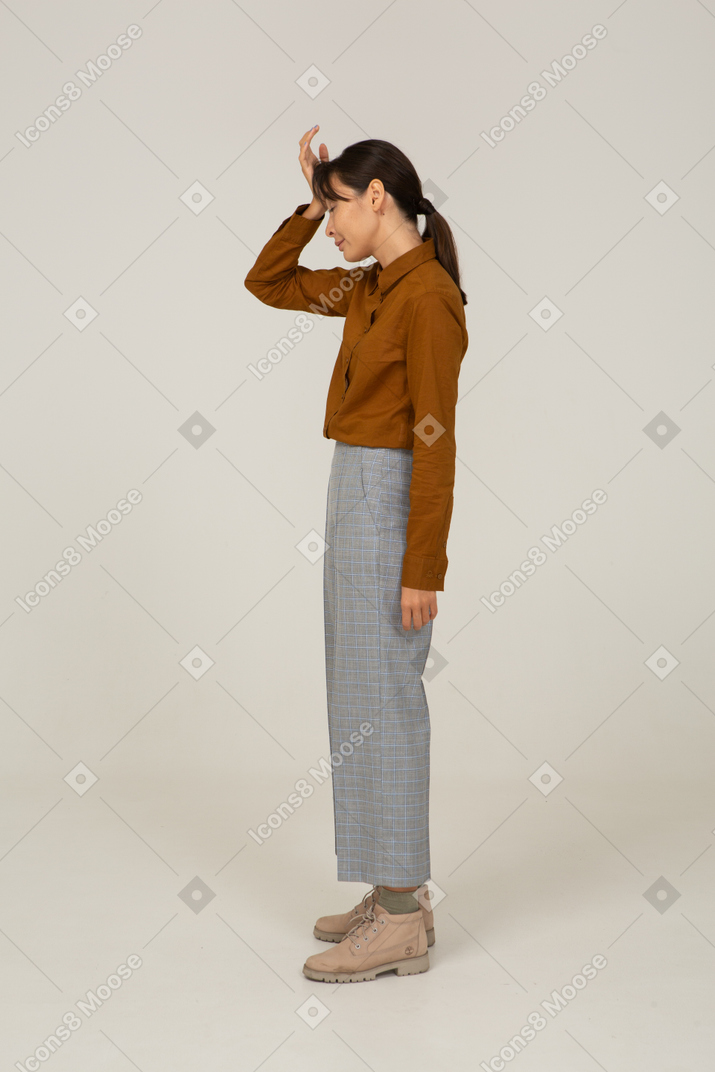 Vista laterale di una giovane donna asiatica in calzoni e camicetta che tocca la testa