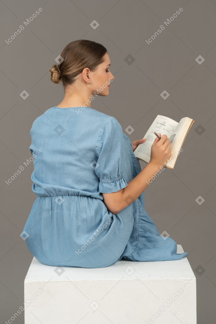 Side-back view of young woman sitting on a cube and writing in notebook