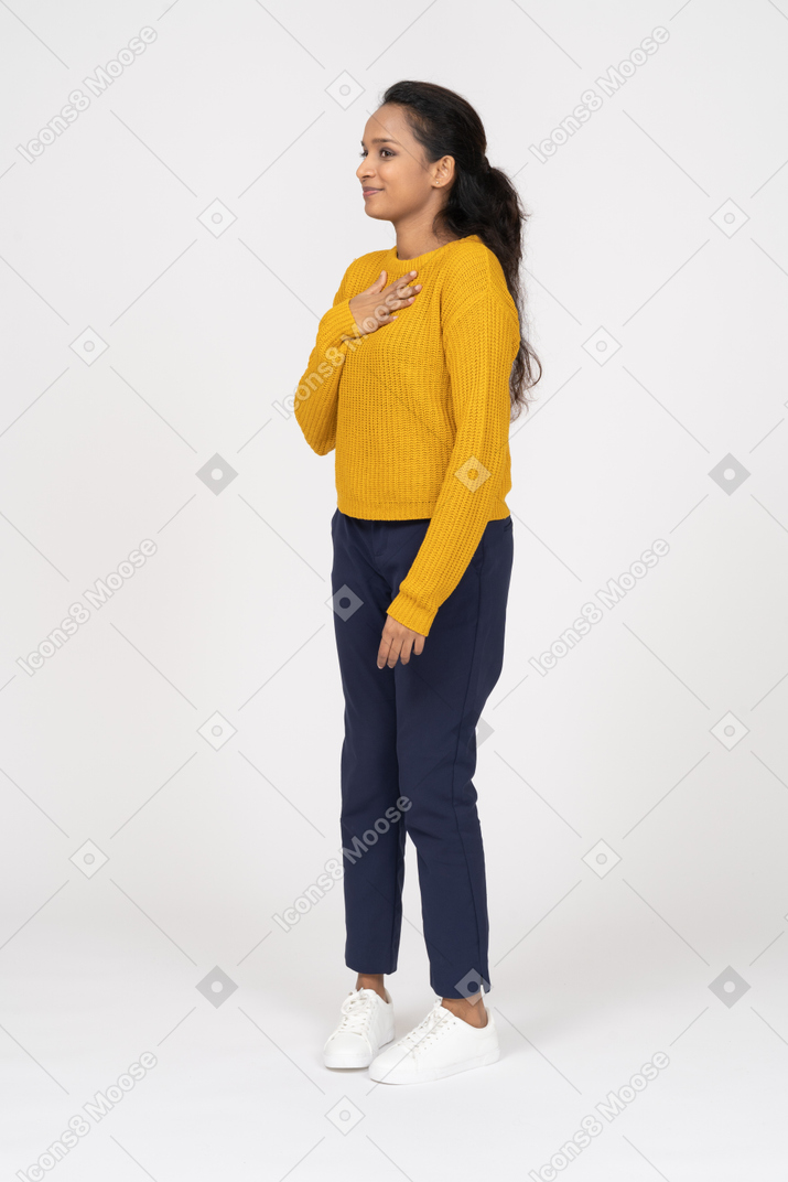 Vista lateral de una niña feliz en ropa casual posando con la mano en el pecho