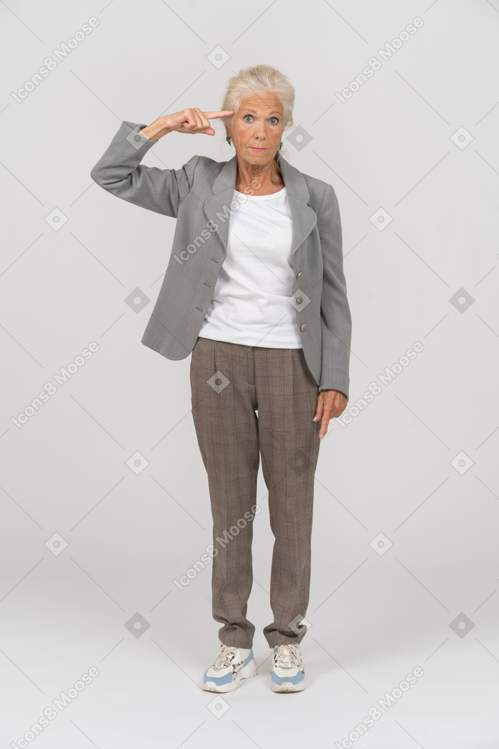 Vista frontal de una anciana en traje mirando a la cámara y mostrando el signo de tornillo suelto