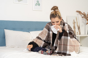 Front view of an ill young lady in pajamas wrapped in checked blanket checking her temperature