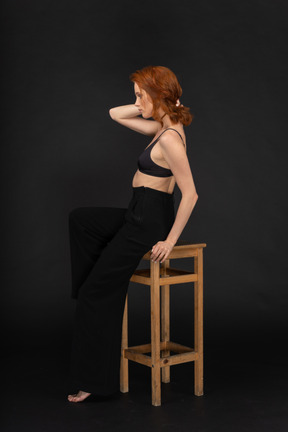 A side view of the young beautiful woman dressed in black pants and bra, sitting on the wooden chair and holding her hand on the hair