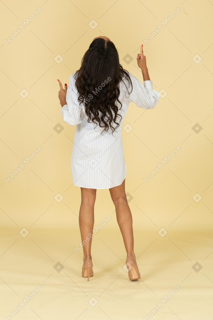 Vista traseira de uma jovem mulher de pele escura dançando em seu vestido branco levantando as mãos
