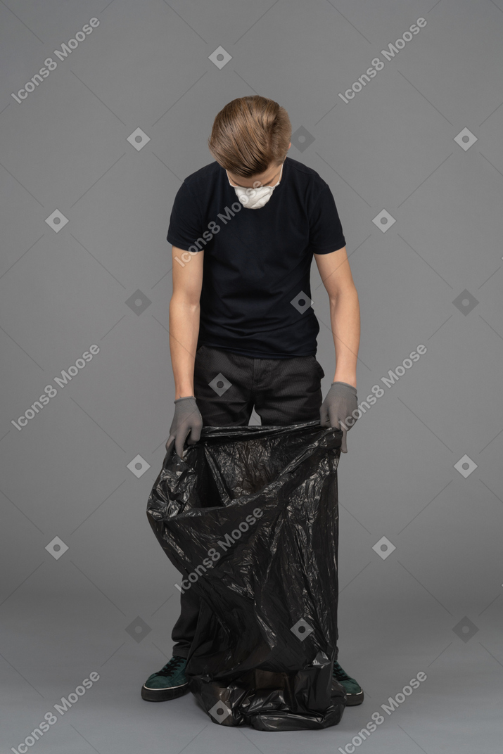 A young man wearing a mask and looking inside a trash bag