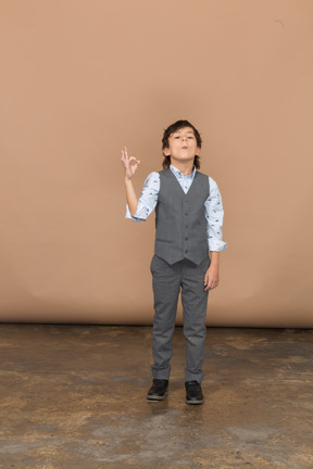 Vista frontal de um menino bonito de terno cinza mostrando sinal de ok