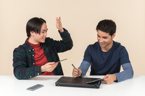 Two smiling young geeks sitting at the table and fixing laptop