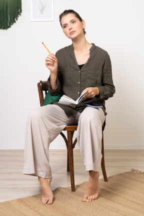 一个体贴的年轻女子穿着家居服坐在椅子上，拿着铅笔和笔记本的前视图
