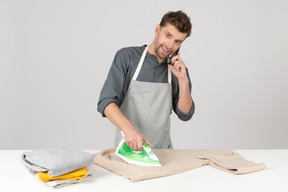 Young househusband ironing clothes and talking on the phone