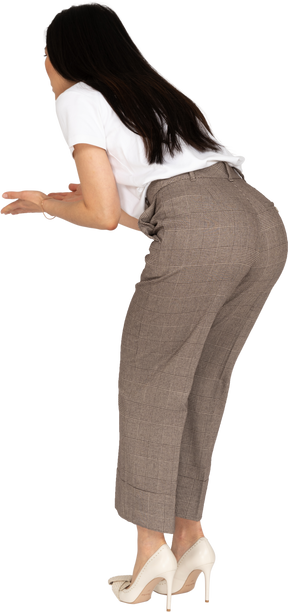 Vista posteriore di tre quarti di una giovane donna interrogativa in calzoni e maglietta che alza le mani e si china