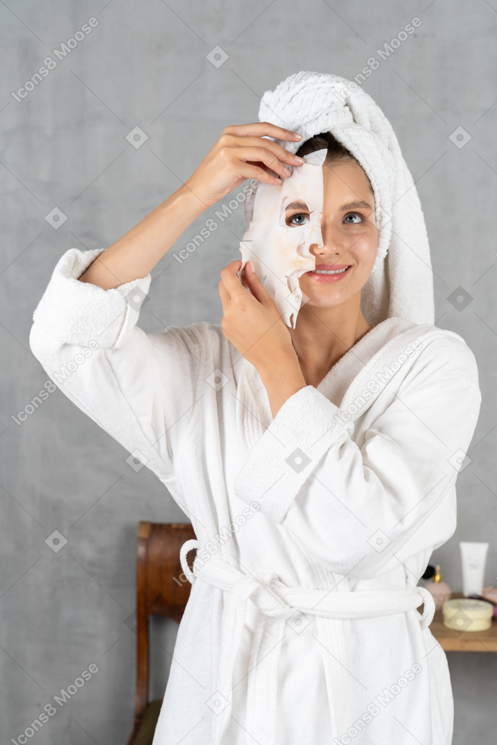 Woman in bathrobe holding a sheet mask over half of her face