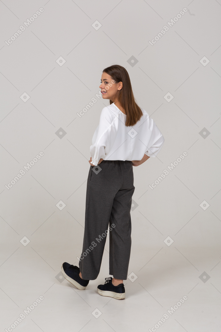 腰に手を置いているオフィスの服を着た傲慢な若い女性の4分の3の背面図