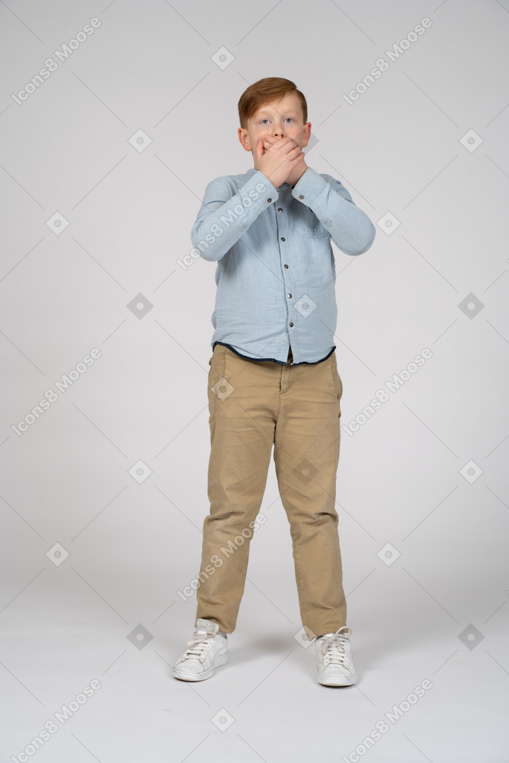 Front view of a scared boy covering mouth with hands and looking at camera