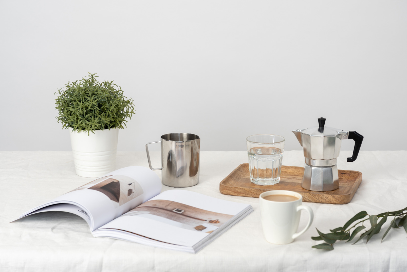 Moka and glass of water on the wooden tray, plant in pot, magazine, large cup of milk coffee and pitcher