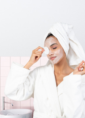 A woman in a bathrobe wiping off a face mask