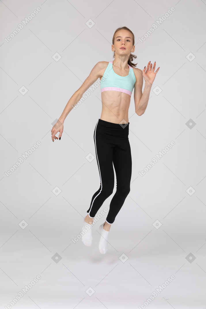 Vista de três quartos de uma adolescente em roupas esportivas levantando a mão e pulando