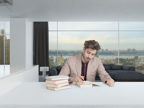 Красивый молодой человек в бежевом пиджаке сидит за столом в просторной современной комнате, читая книги