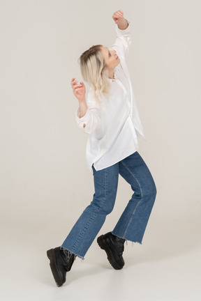 Vista di tre quarti di una donna bionda in abiti casual che balla in punta di piedi e alza le mani e la testa