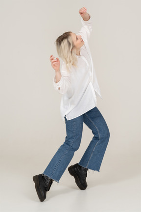 Three-quarter view of a blonde female in casual clothes dancing on her tiptoes and raising hands and head