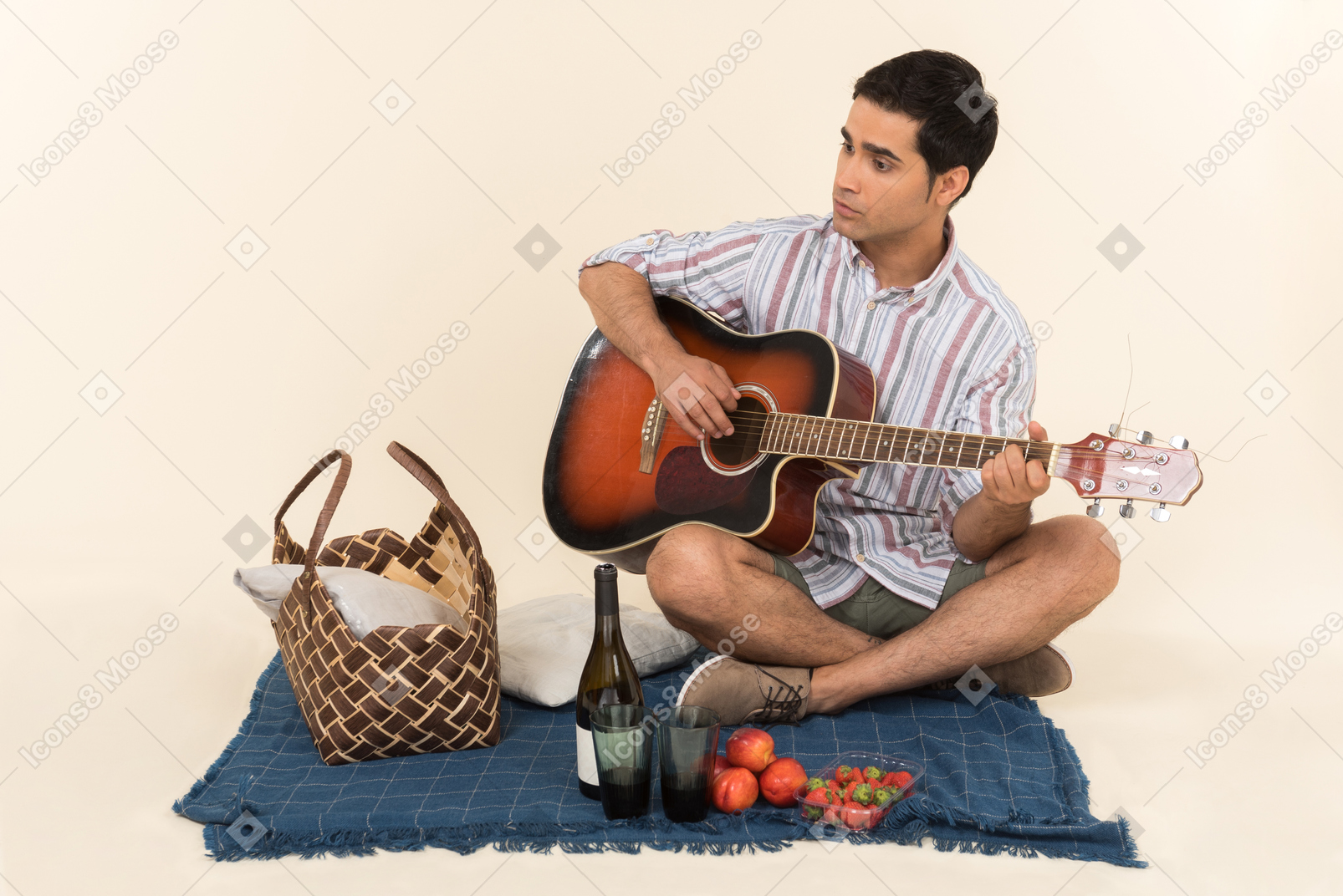 Junger kaukasischer kerl, der nahe picknickkorb auf der decke sitzt und gitarre spielt