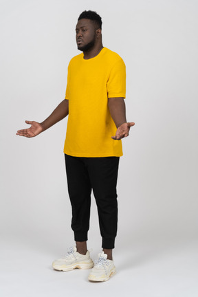 Vista de três quartos de um homem jovem de pele escura descontente em uma camiseta amarela estendendo as mãos