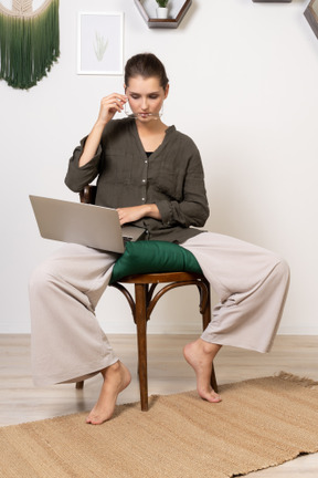 Vista frontal de una mujer joven vestida con ropa de casa sentada en una silla con una computadora portátil y poniéndose gafas