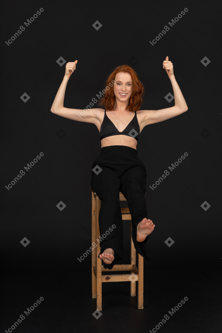 A frontal view of the beautiful woman dressed in black pants and bra, sitting on the wooden chair, smiling and looking to the camera