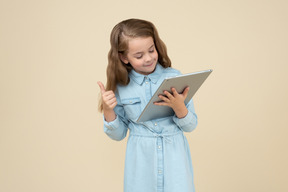 Cute little girl holding a tablet and showing thumbs up