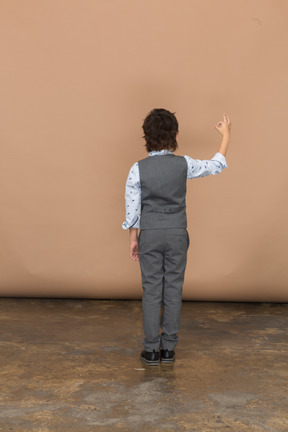 Vista trasera de un niño con traje gris que muestra el signo de ok