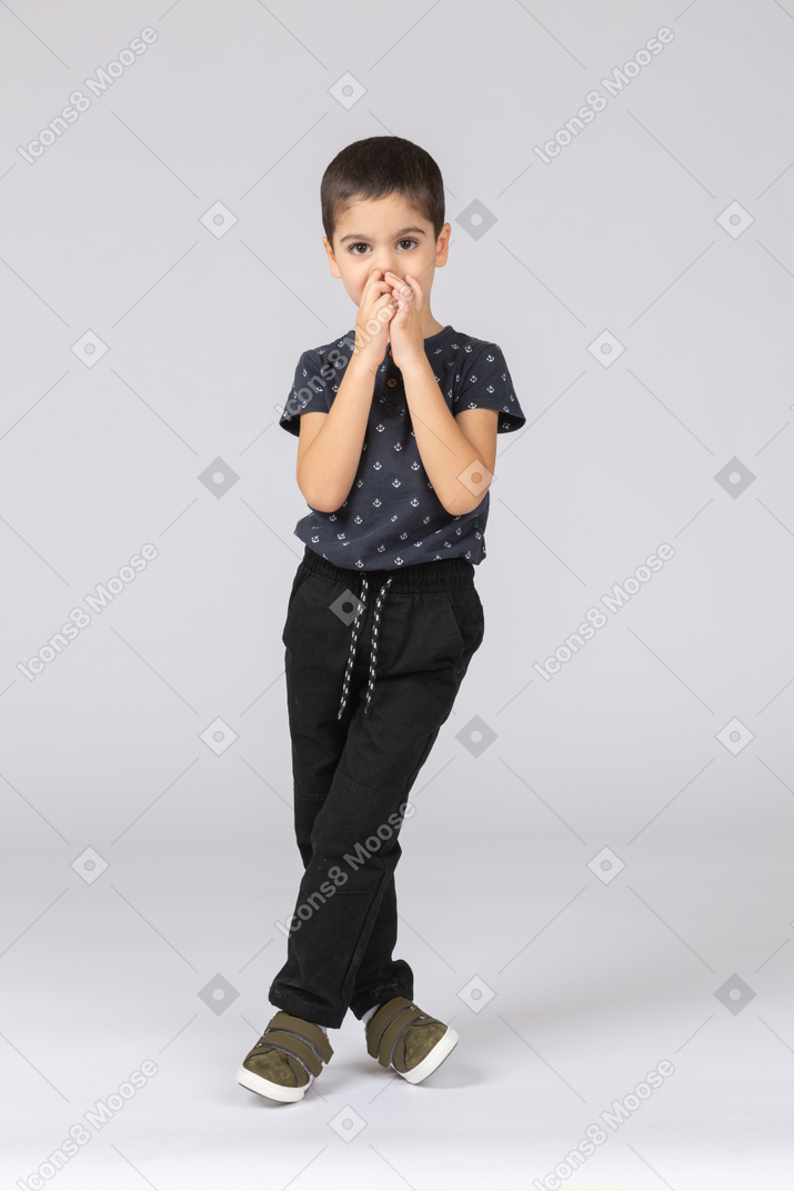 Front view of a cute boy standing with crossed legs and touching nose