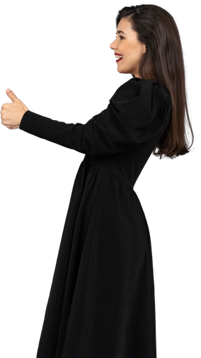Seitenansicht einer lächelnden jungen dame in einem schwarzen kleid, das einen daumen nach oben zeigt