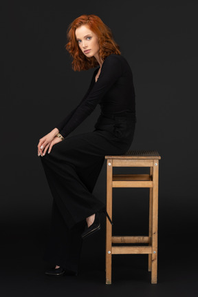 A side view of the sexy woman sitting on the tall wooden chair and looking to the camera