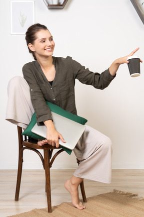 Vista frontal de uma jovem sorridente, sentada em uma cadeira, segurando seu laptop e a xícara de café