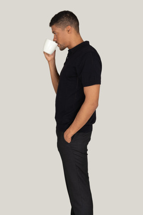 Standing in profile young man in black t-shirt and pants drinking coffee