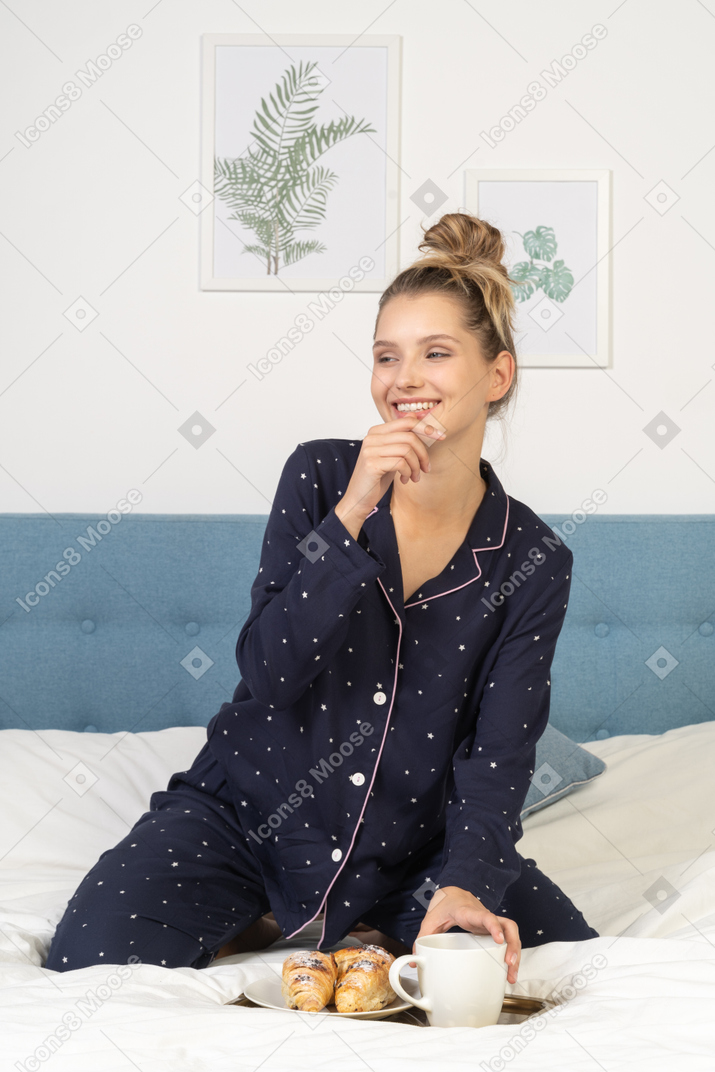 Front view of a young lady in pajamas holding a cup of coffee and some pastries sitting in bed