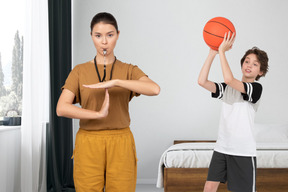 A female coach showing time out hand gesture next to a boy with basketball ball in a bedroom