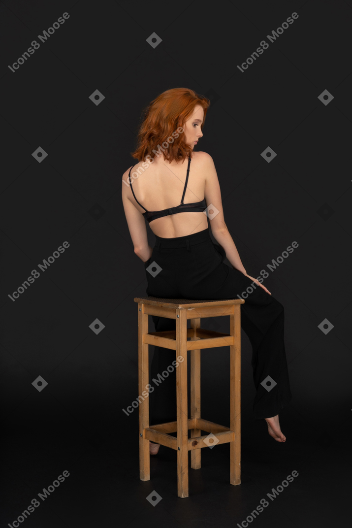 A back side view of the beautiful woman dressed in black pants and bra, sitting on the wooden chair, holding her hand on the knee and looking down