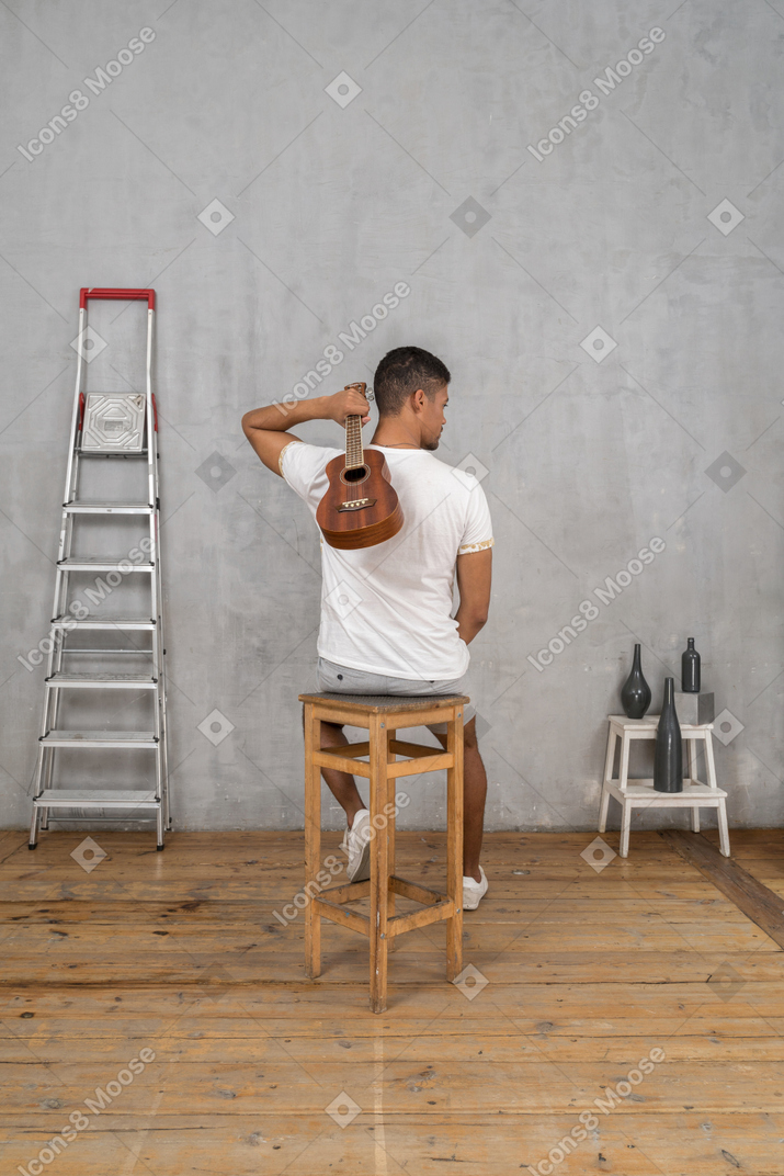 Vista posteriore di un uomo su uno sgabello che tiene un ukulele dietro la schiena con una mano