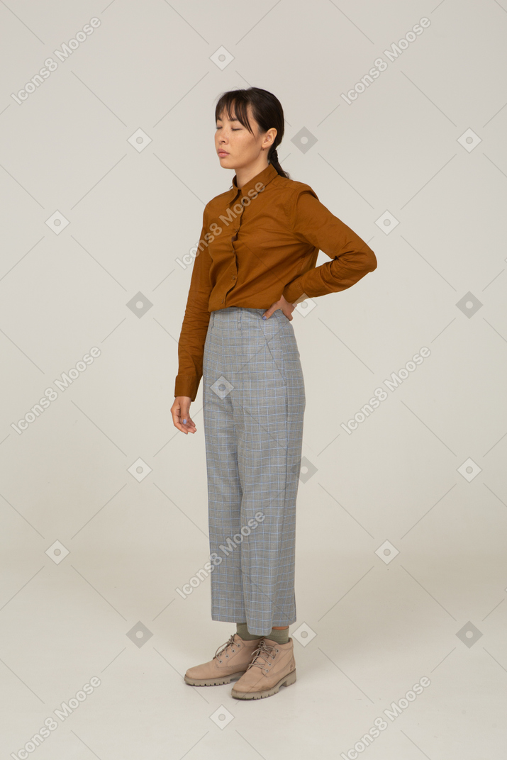Vista di tre quarti di una giovane donna asiatica assonnata in calzoni e camicetta in piedi e mettendo la mano sull'anca